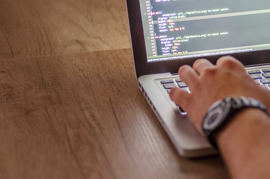 A close-up view of a person's hand typing on a silver laptop keyboard placed on a wooden table. The laptop screen displays lines of colorful code against a dark background, indicating a programming or