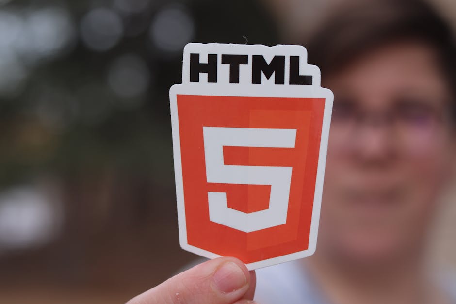 A person holding a sign with the HTML5 logo, featuring a white shield with an orange background and the text 'HTML5' at the top. The background is blurred, showing hints of greenery and trees, suggest