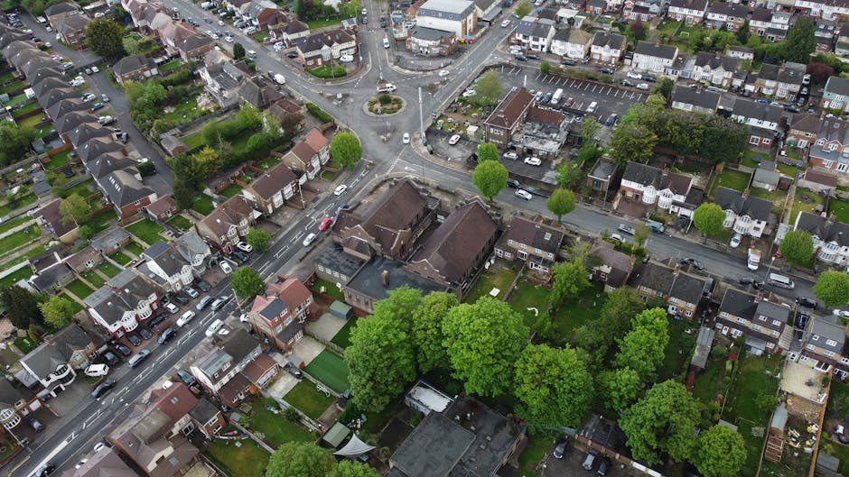 An aerial view of a residential neighbourhood with a variety of small front gardens, neatly trimmed lawns, and lush green trees lining the streets. The image shows a mix of single and semi-detached ho