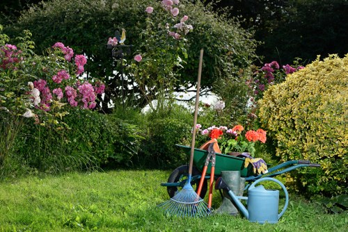 A well-maintained garden scene featuring a lush, green lawn bordered by vibrant flowering shrubs and bushes, including pink and purple roses and other colorful blooms. The garden is enclosed by tall, leafy trees and dense greenery in the background, creating a sheltered outdoor space. In the foreground, various gardening tools and equipment are arranged on the grass, including a blue watering can, a garden rake, gloves, a small garden fork, and a wheelbarrow filled with additional tools and supplies, indicating ongoing yard work or maintenance. The scene appears to be outdoors on a bright, slightly overcast day, with natural light highlighting the rich textures and natural tones of the plants and garden materials, reflecting a tidy, cared-for garden environment typical of Kenton or nearby areas. The setting emphasizes outdoor gardening and lawn care services provided by Gardening Services Kenton, suitable for transforming residential gardens into beautiful, manageable outdoor spaces.